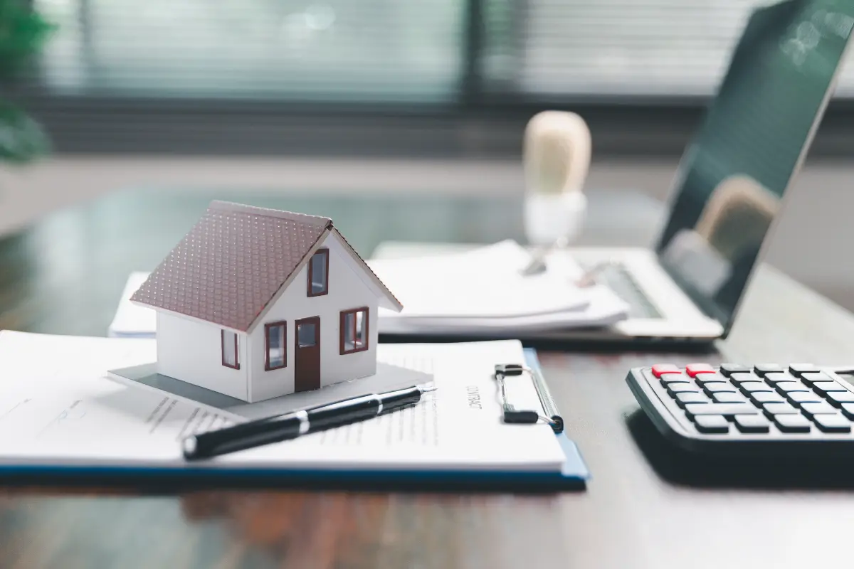 Miniature house model placed on mortgage documents with a pen, laptop, and calculator on a desk, representing home loans and financial planning.